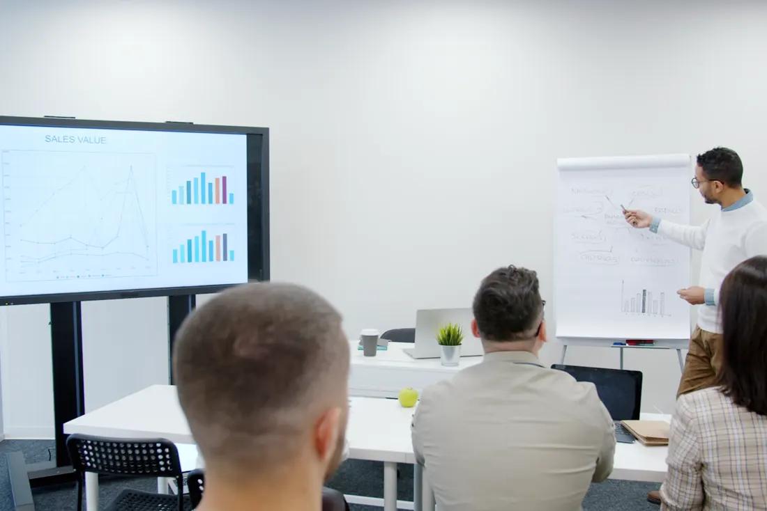 Formateur au paperboard devant un petit groupe assis, écran de présentation à côté de lui